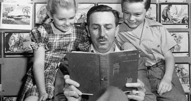Black and white photo of Walt Disney reading a book to two children, with a backdrop of illustrated storyboards, capturing a moment of storytelling and inspiration.